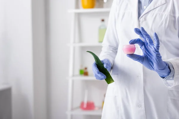 Vue recadrée de l'assistant de laboratoire dans des gants en latex tenant la feuille d'aloès et le récipient avec crème — Photo de stock