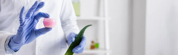 Cropped view of laboratory assistant in latex gloves holding aloe leaf and container with cream, banner — Stock Photo