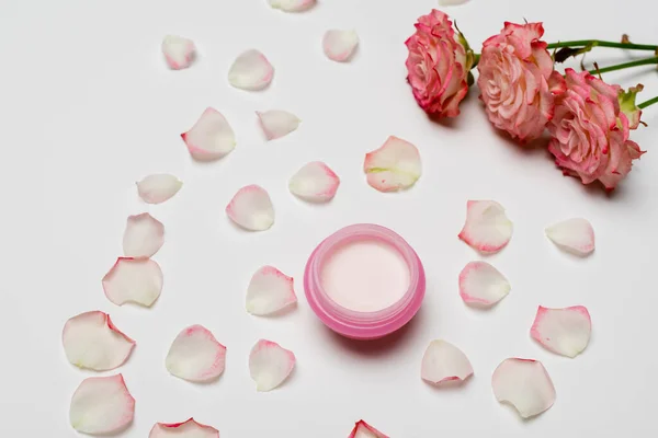 Vista de ángulo alto de flores y pétalos de color rosa cerca de un recipiente pequeño con crema en blanco — Stock Photo