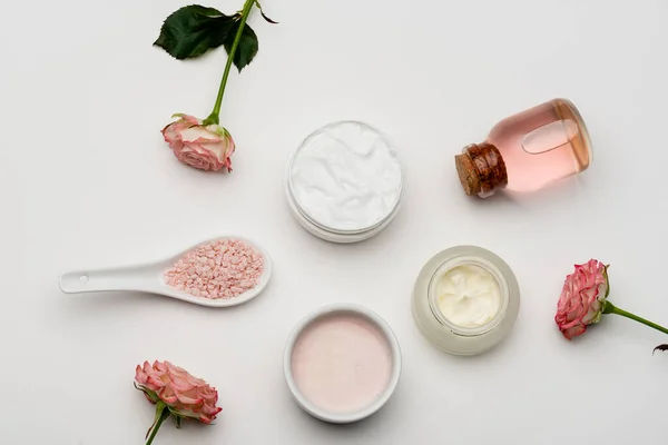 Vista superior de flores perto de sal marinho rosa, garrafa com água de rosas e recipientes com creme em branco — Fotografia de Stock