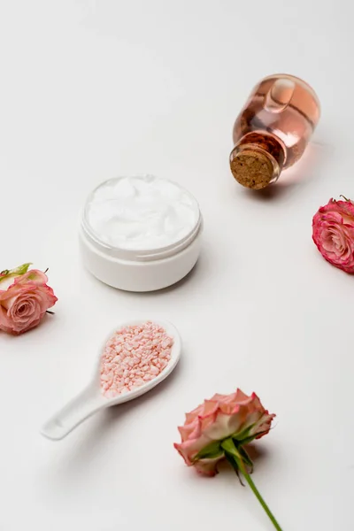 Blüten in der Nähe von rosa Meersalz, Flasche mit Rosenwasser und Behälter mit Sahne auf weiß — Stockfoto