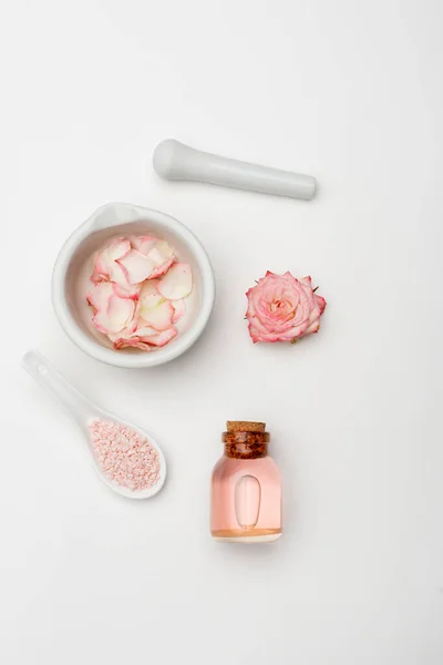 Top view flower near pestle and mortar with petals, sea salt and bottle with rose water on white — Stock Photo