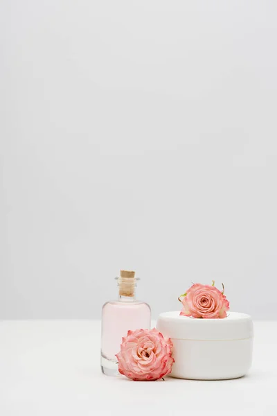 Bottle with extract near container with cream and pink flowers on white — Stock Photo