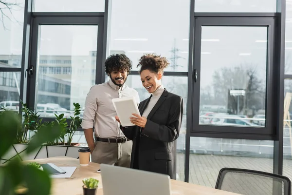 Sonriente mujer de negocios afroamericana sosteniendo tableta digital cerca de colega indio en la oficina - foto de stock