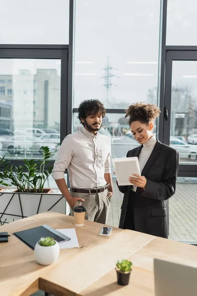 Joven mujer de negocios afroamericana usando tableta digital cerca de colega indio y dispositivos en la oficina - foto de stock