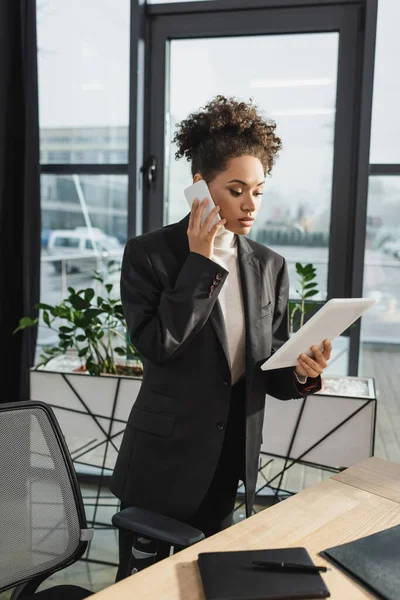 Empresaria afroamericana hablando por celular y sosteniendo tableta digital en la oficina - foto de stock