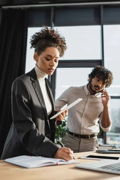 Afroamerikanische Geschäftsfrau hält digitales Tablet in der Hand und schreibt auf Notizbuch nahe indischer Kollegin, die im Büro mit dem Handy spricht — Stockfoto