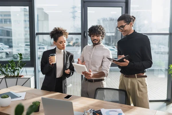 Empresario indio sosteniendo tableta digital cerca de colegas afroamericanos con café y portátil en la oficina - foto de stock