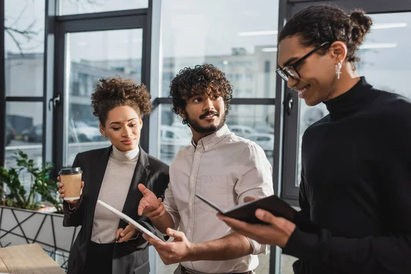 Hombre de negocios indio sonriente apuntando a la tableta digital cerca de colegas afroamericanos en el cargo - foto de stock