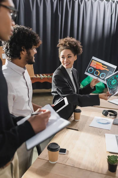 Smiling african american businesswoman holding papers near interracial colleagues with notebook and digital tablet — Stock Photo