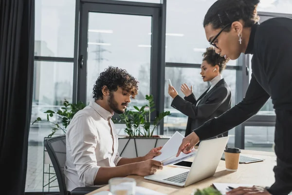 Vista lateral del hombre de negocios afroamericano apuntando a los periódicos cerca de colega indio y dispositivos en la oficina - foto de stock