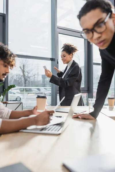 Smiling african american businesswoman having video call on smartphone near blurred multiethnic colleagues in office — Stock Photo