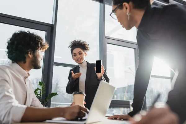 Smiling african american businesswoman pointing at cellphone near blurred multiethnic colleagues in office — Stock Photo