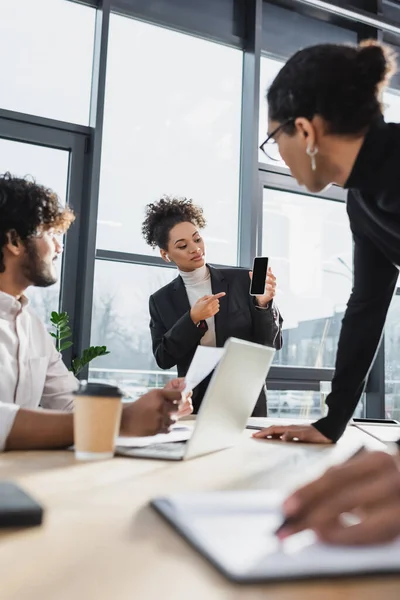 Jovem empresária afro-americana apontando para smartphone perto de empresários multiétnicos no escritório — Fotografia de Stock