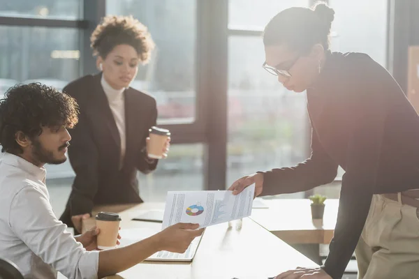 Interracial businessmen holding document near laptop and blurred african american colleague in office — Stock Photo