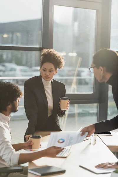 African american businesswoman holding coffee to go near blurred colleagues with paper in office — Stock Photo