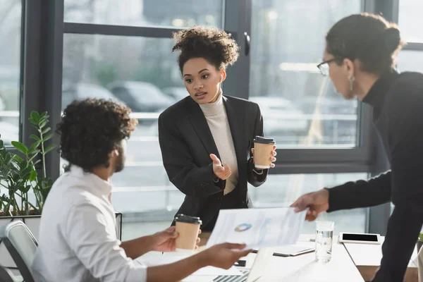 Empresaria afroamericana sosteniendo café mientras habla con colegas multiétnicos con papel en la oficina - foto de stock