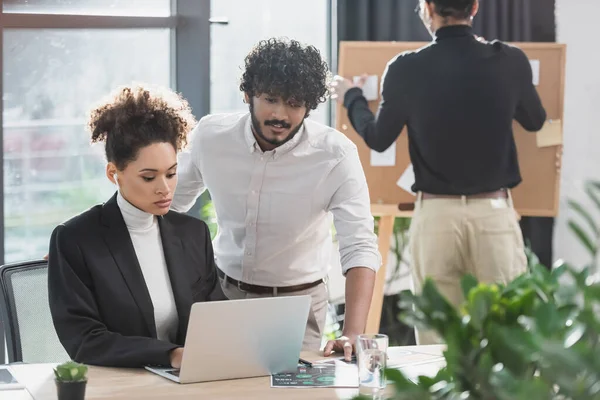 Indian businessman standing near african american colleague using laptop in office — Stock Photo