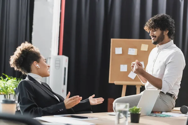 Vista lateral de la alegre mujer de negocios afroamericana en auriculares hablando con un colega indio cerca de gadgets en la oficina - foto de stock