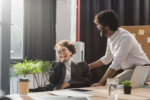 Laughing interracial colleagues having fun near devices in office — Stock Photo