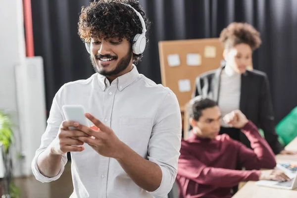 Uomo d'affari indiano sorridente in cuffia utilizzando smartphone in ufficio — Foto stock