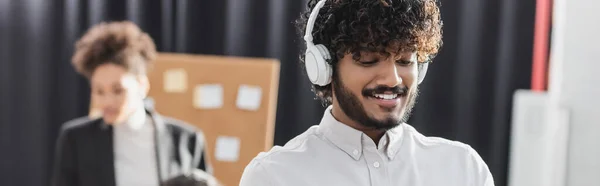 Empresário indiano positivo em fones de ouvido no escritório, banner — Fotografia de Stock