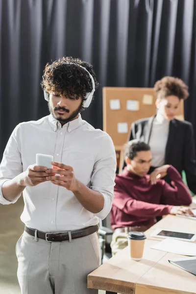 Empresario indio en auriculares con teléfono inteligente cerca borrosa colegas afroamericanos en la oficina - foto de stock