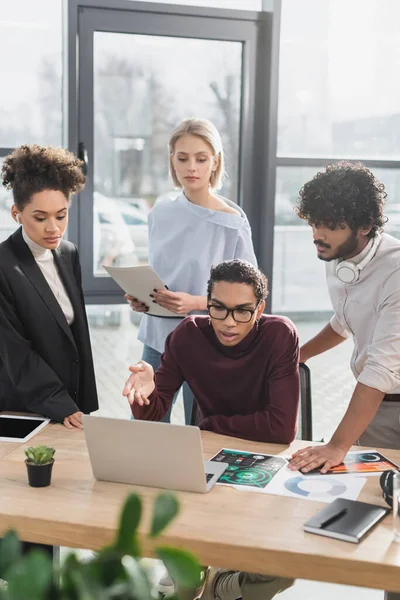 Multikulturelle Geschäftsleute schauen im Büro auf Laptop in der Nähe von Zeitungen — Stockfoto