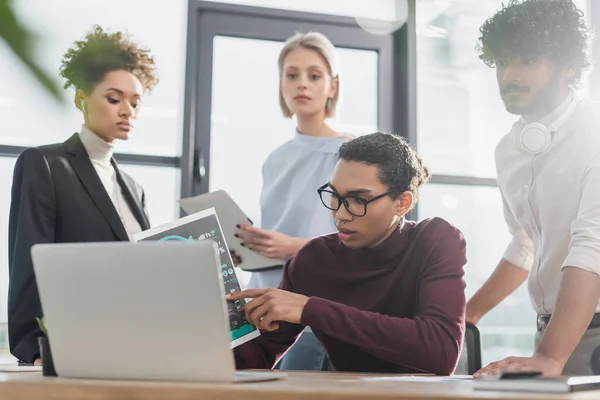 Afrikanischer Geschäftsmann zeigt auf Dokument in der Nähe von Laptop und Bürokollegen — Stockfoto