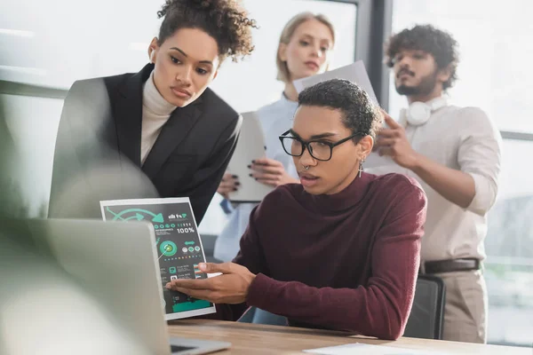 Empresario afroamericano señalando el papel durante una videollamada en una computadora portátil cerca de colegas multiétnicos borrosos en la oficina - foto de stock