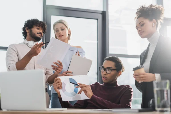 Empresarios afroamericanos con café y papel con videollamada en laptop cerca de colegas multiétnicos en la oficina - foto de stock