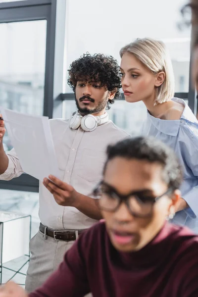 Junge multiethnische Geschäftsleute betrachten Papier in der Nähe eines verschwommenen afrikanisch-amerikanischen Kollegen im Büro — Stockfoto