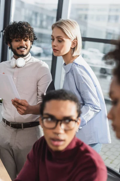 Interracial business people working with document near blurred african american colleagues in office — Stock Photo