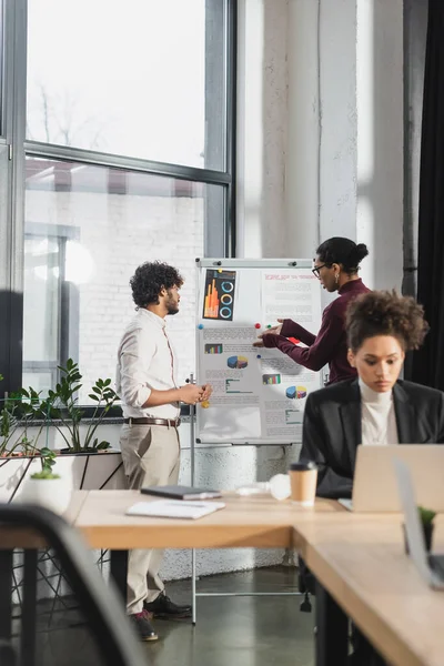 African american businessman pointing at papers on flip chart near indian colleague in office — Stock Photo