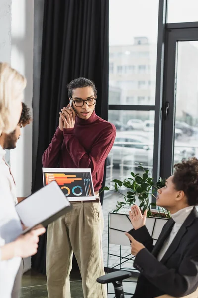 Afrikanischer Geschäftsmann spricht im Büro mit Smartphone neben Kollegen mit Papier und Notizbuch — Stockfoto