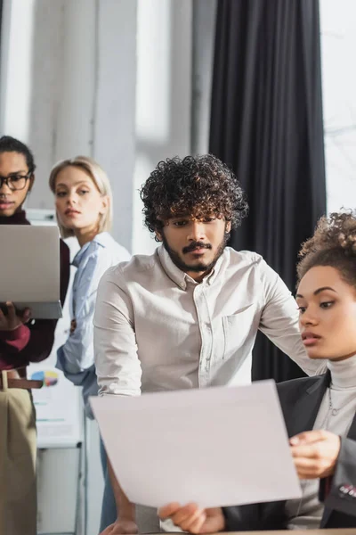 Indian businessman looking at blurred document near african american colleague in office — Stock Photo