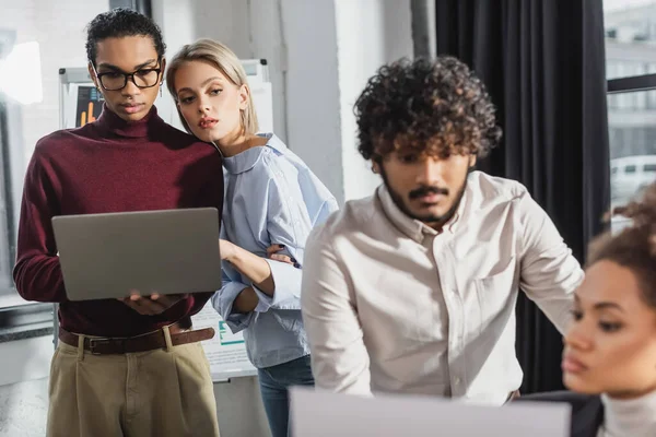 Giovane donna d'affari che utilizza laptop con collega afro-americano vicino a uomini d'affari in ufficio — Foto stock