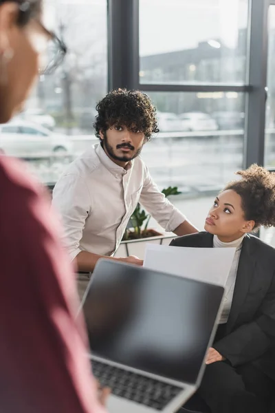 Uomo d'affari indiano guardando collega offuscata con computer portatile vicino afro-americano donna d'affari con carta in carica — Foto stock