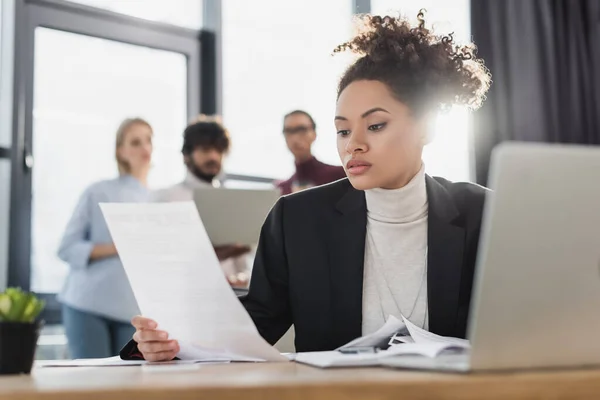 Empresaria afroamericana sosteniendo papel cerca de computadora portátil borrosa en la oficina - foto de stock