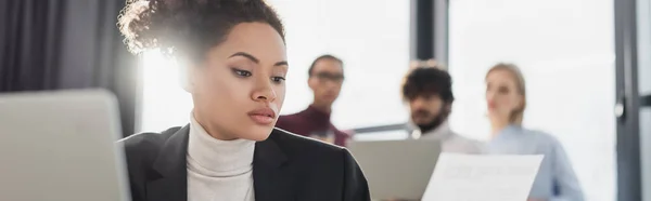 Молода афроамериканська бізнесменка, яка дивиться на папір біля розмитого ноутбука і колег по роботі, банер. — стокове фото