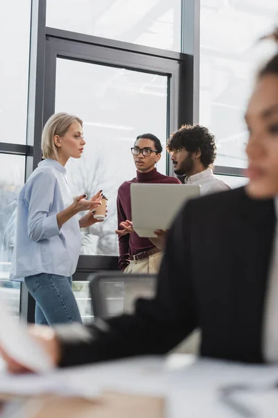 Empresaria sosteniendo café y hablando con colegas interracial con portátil en la oficina - foto de stock