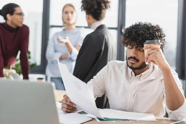 Uomo d'affari indiano stanco che tiene caffè e documento vicino al computer portatile in ufficio — Foto stock