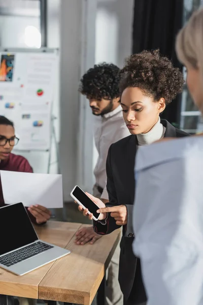 Femme d'affaires afro-américaine pointant vers smartphone près de collègues multiethniques au bureau — Photo de stock