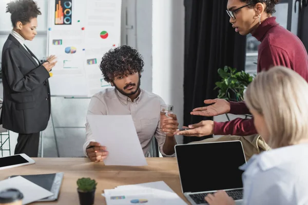 Indian businessman holding paper and water near interracial colleagues in office — Stock Photo