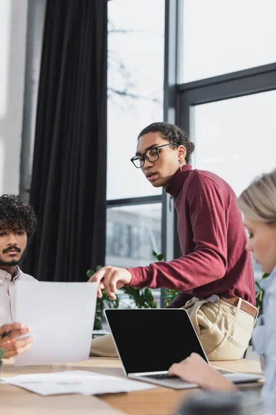 Empresario afroamericano hablando con colegas multiétnicos con papel y portátil en la oficina — Stock Photo