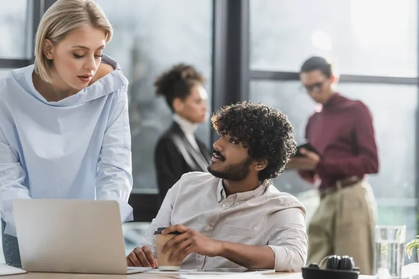 Empresária conversando com colega indiano com café para ir perto de laptop no escritório — Fotografia de Stock