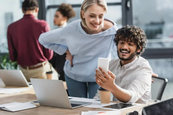 Smartphone in der Hand eines verschwommenen indischen Geschäftsmannes, der in der Nähe seines Bürokollegen arbeitet — Stockfoto