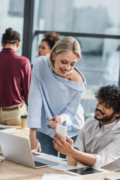 Lächelnde Geschäftsfrau zeigt auf Smartphone neben indischem Kollegen und Geräten im Büro — Stockfoto