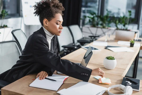 Giovane donna d'affari afro-americana che prende appunti appiccicosi vicino al laptop in ufficio — Foto stock