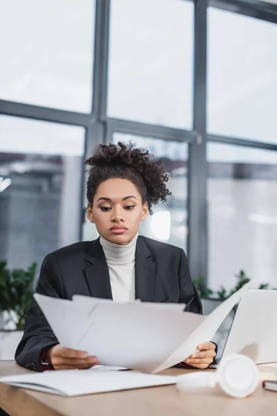 Empresaria afroamericana sosteniendo papeles cerca de laptop en oficina - foto de stock